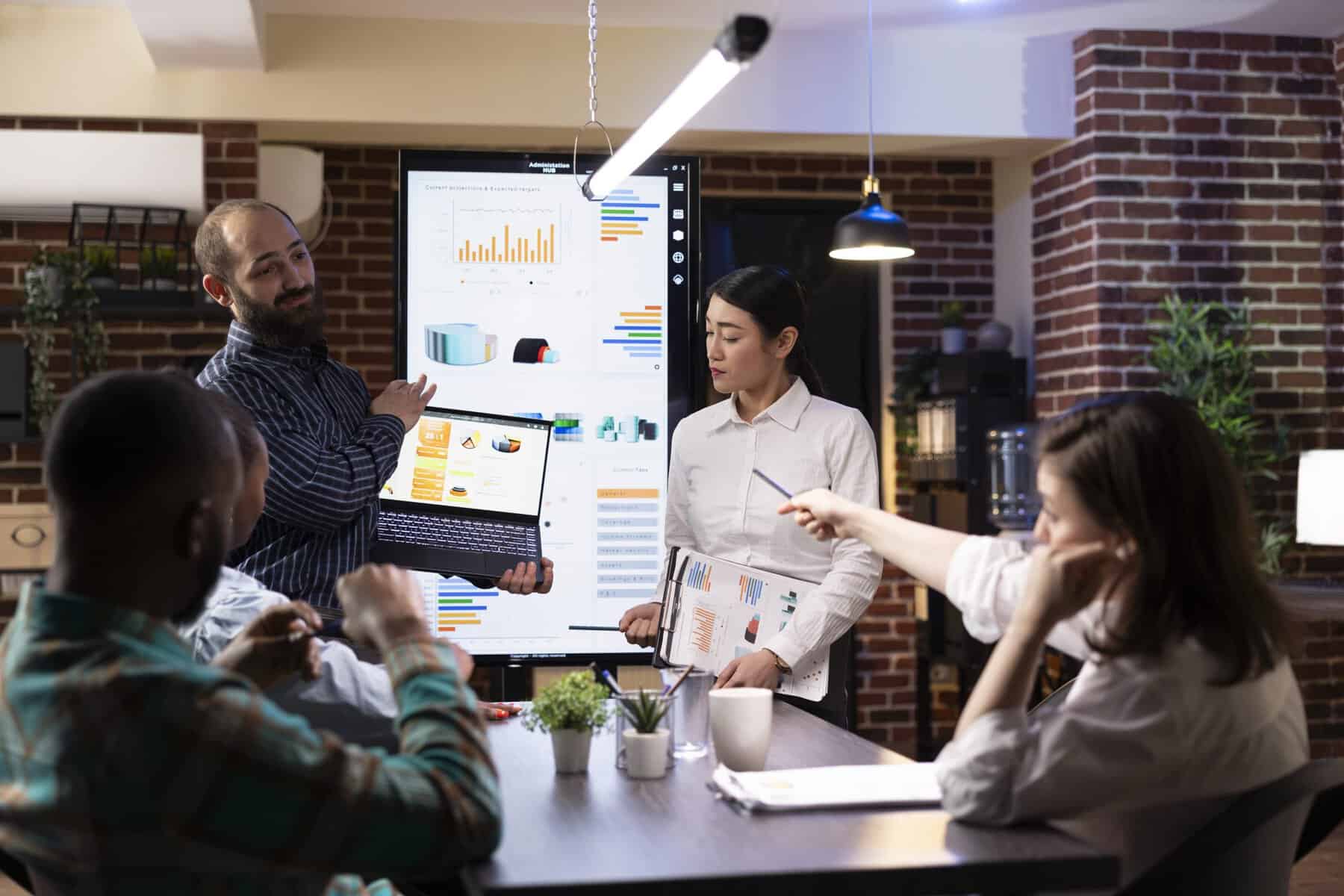 Male and female professionals present financial charts on clipboard and digital devices, engaging their team in insightful business discussion. Diverse colleagues gathered for late night meeting.
